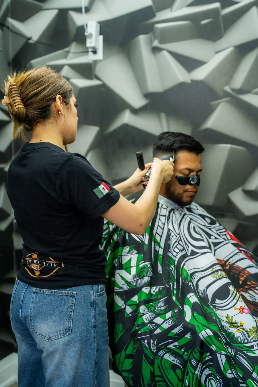 Barber cutting a client in a patterned cape, standing, textured wall