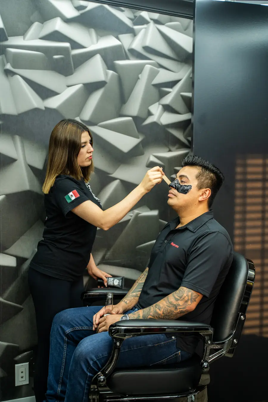 Barber applying a charcoal face mask to a seated client, textured wall